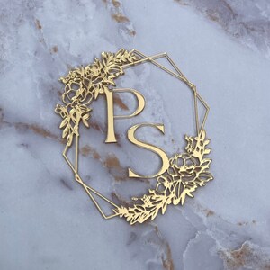 May include: Gold metal monogram featuring the letters "P" and "S" encircled by a floral design. The monogram is displayed against a white marble backdrop.