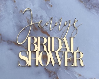 May include: Gold glitter letters spelling "Jenny's Bridal Shower" on a white marble background.