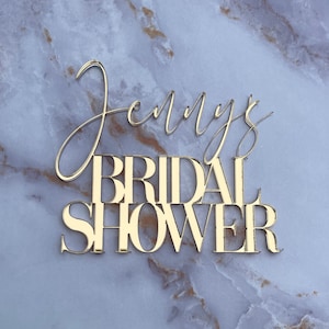 May include: Gold glitter letters spelling "Jenny's Bridal Shower" on a white marble background.
