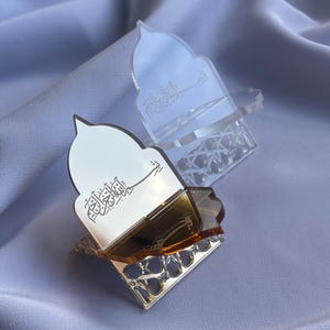 Könnte beinhalten: Zwei dekorative Halter aus Gold und klarem Acryl mit arabischer Kalligraphie. Die Halter haben eine gewölbte Oberseite und eine geometrische Basis. Einer ist goldfarben mit klarem Oberteil, der andere ist klar.