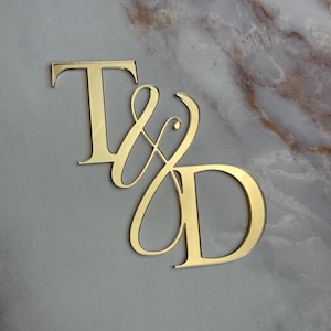 May include: Gold mirror acrylic letters spelling "T & D" in a cursive font. The letters are on a gray marble background.