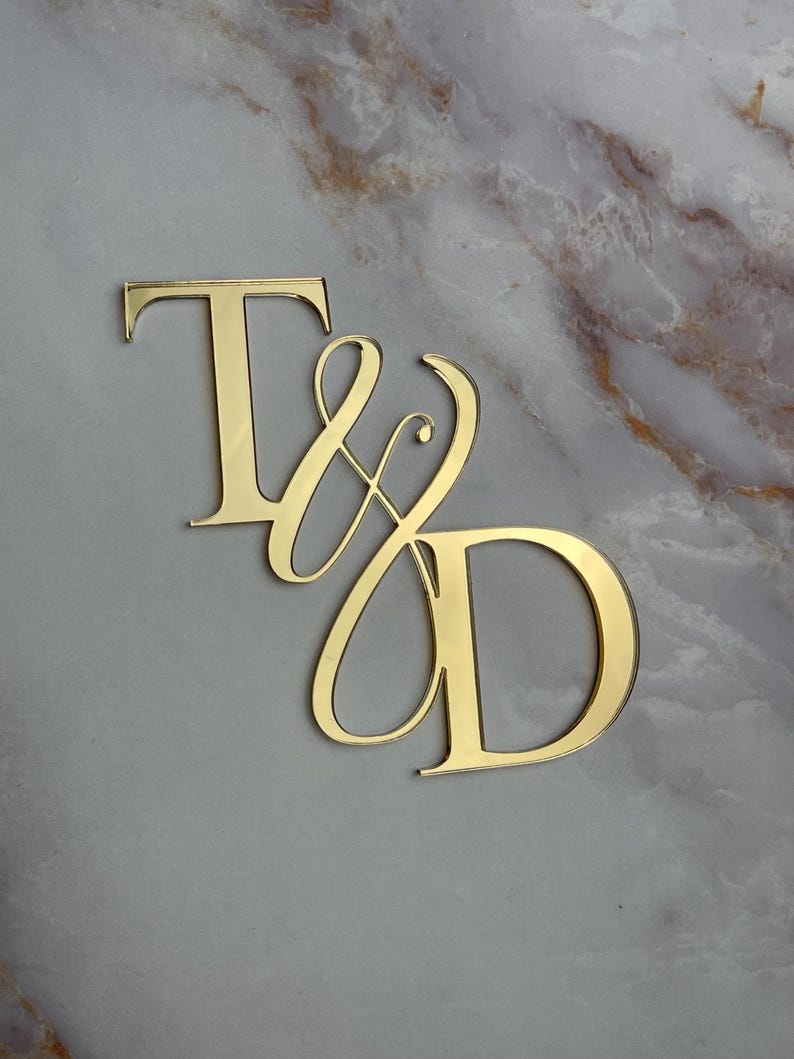 May include: Gold mirror acrylic letters spelling "T & D" in a cursive font. The letters are on a gray marble background.