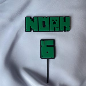 Peut inclure: Décorations de gâteau vertes et noires avec le nom "NOAH" et le chiffre "6" dans une police pixélisée. Le dessus de gâteau avec le nom est rectangulaire, et celui avec le chiffre a un bâton noir.