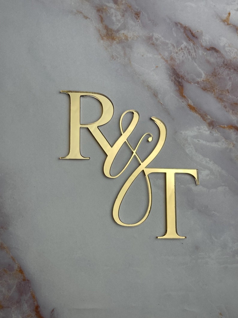 May include: Gold-colored acrylic letters spelling "R & T" with a decorative script font for the ampersand. The letters are on a marble background.