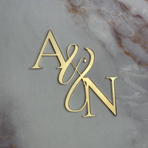May include: Gold-colored metal initials "A & N" with a decorative script font. The letters are cut out and stand on a marble surface.