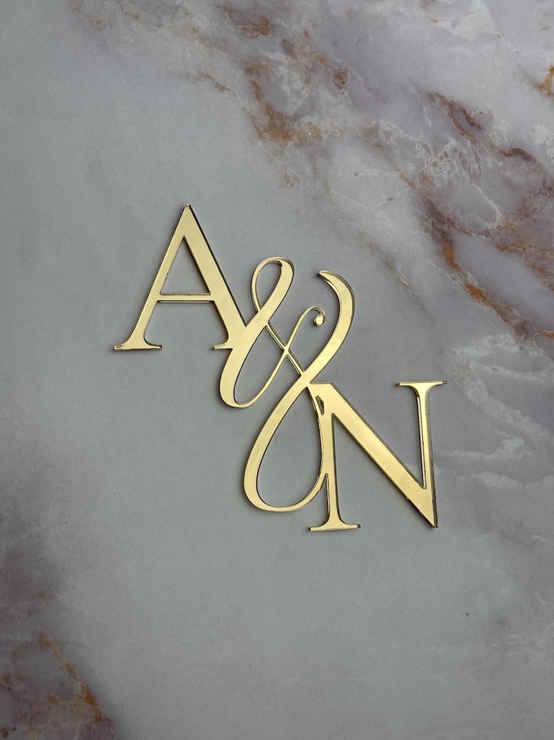 May include: Gold-colored metal initials "A & N" with a decorative script font. The letters are cut out and stand on a marble surface.