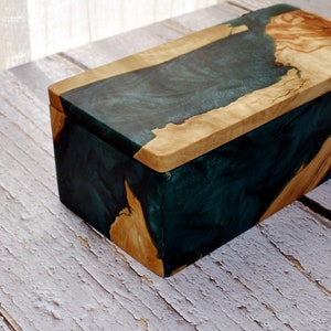 Puede incluir: Una caja de madera con una incrustación de resina verde oscuro. La tapa es de madera y tiene un acabado de madera natural.