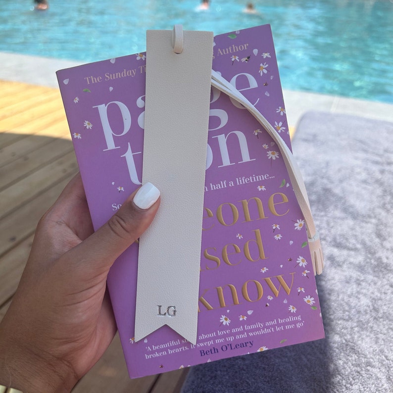 White Vegan Leather Bookmark with pointed arrow ends and detachable tassel with LG printed in silver, staged with a book by the pool