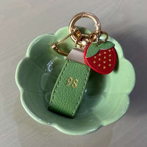May include: A green leather keychain with the number 98 in gold lettering, a gold-colored clasp, and a red strawberry charm with green leaves and gold seeds. The keychain rests in a light green, scalloped-edge dish.