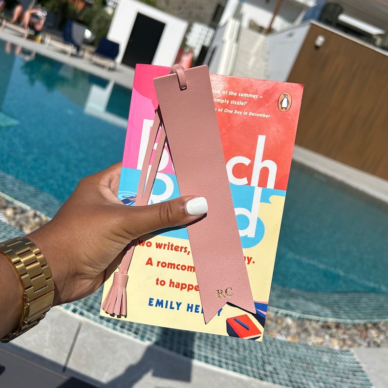 Pink Vegan Leather Bookmark with pointed arrow ends and detachable tassel with RC printed in gold, staged with a book by the pool