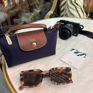 May include: A navy blue handbag with a brown leather flap and handle, featuring the initials "LG". Tortoiseshell sunglasses and a black camera are also present. A white card holder with "LG" is next to the sunglasses.