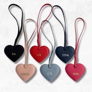 May include: A collection of heart-shaped luggage tags in various colors, including black, red, pink, and blue. Each tag features a gold-colored initial or name. The tags have a matching strap for attachment.
