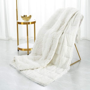 May include: A white fluffy throw blanket with a quilted pattern. The blanket is draped over a chair and a small gold side table.