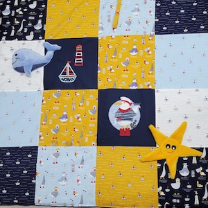 Könnte beinhalten: Eine blaue und gelbe Patchwork-Spielmatte mit verschiedenen Applikationen zum Thema Meer, darunter ein Wal, ein Leuchtturm, ein Segelboot und eine Möwe. Die Matte ist perfekt für Babys und Kleinkinder zum Spielen.