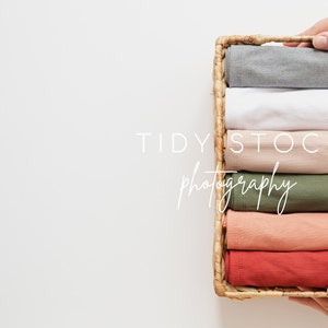 May include: A wicker basket filled with six folded shirts in various colors, including gray, white, beige, green, peach, and red. The basket is being held by two hands.