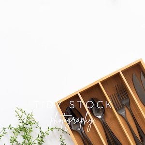 Peut inclure: Un plateau en bois pour couverts avec des ustensiles en métal noir, y compris des cuillères, des fourchettes et un couteau. Le plateau est sur une surface blanche avec une plante verte dans le coin inférieur gauche.