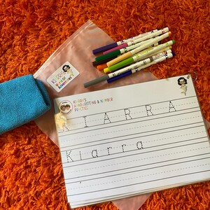 May include: A handwriting practice workbook with the name "Kiarra" printed on the pages, accompanied by a set of colorful markers and a small blue cloth. The workbook is designed for handwriting and number practice.