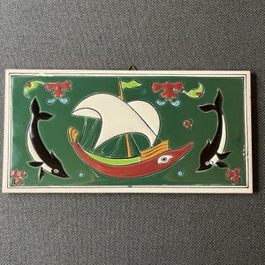May include: A rectangular ceramic wall tile featuring a dark green background with a central red and yellow boat with white sails. Two black and white dolphins are positioned on either side of the boat, accompanied by red floral motifs.