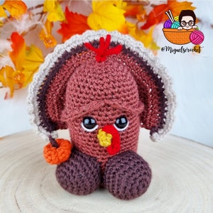 Op de afbeelding: Een gehaakt amigurumi-speelgoed in de vorm van een kalkoen met een rode keellap, een bruin lichaam en een grijze en bruine gevederde staart. De kalkoen houdt een kleine oranje pompoen vast.