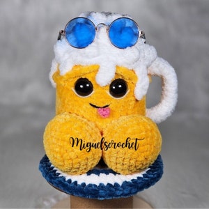 Peut inclure: Un jouet en peluche en forme de mug à bière crocheté avec un dessus en mousse blanche, des lunettes de soleil bleues et un visage souriant. Le mug est posé sur un dessous de verre crocheté bleu et blanc. Le texte "miguelserochet" est visible sur le devant du mug.
