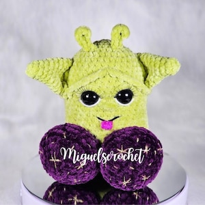 May include: A green crocheted alien plush toy with large black eyes and a pink tongue sticking out. The alien is sitting on two purple crocheted balls with gold glitter.
