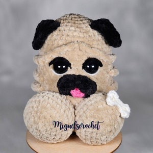 Puede incluir: Un juguete de peluche de pug tejido a crochet con grandes ojos negros, una lengua rosa y un hueso blanco tejido a crochet. El juguete es beige con orejas negras y una nariz negra.