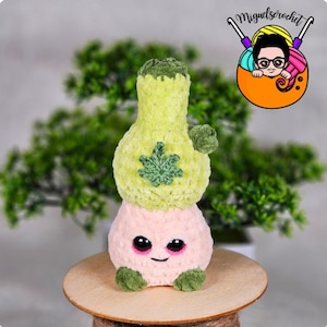 May include: A crocheted plush toy shaped like a cannabis plant. The toy is green and pink and has a smiling face. The toy is sitting on a wooden surface.