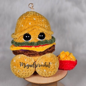 May include: A crocheted plush toy shaped like a hamburger with a red crocheted container of yellow crocheted french fries. The hamburger has a yellow bun, green lettuce, red tomato, brown meat, and purple cheese. The toy has googly eyes and the text "miguelscrochet" is on the front.