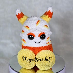 May include: A crocheted candy corn plush toy with orange, yellow, and white stripes. The toy has black eyes and a smiling mouth. The toy is sitting on a white surface with the text "miguelscrochet" visible.
