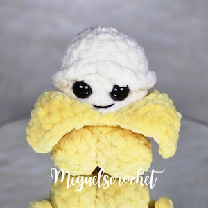 Può includere: Un giocattolo di peluche all'uncinetto a forma di banana con un cono gelato all'uncinetto bianco in cima. Il cono gelato ha occhi neri e una bocca sorridente.