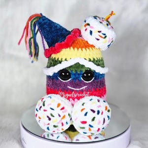 May include: A colorful crocheted birthday cake character with a rainbow body, white frosting, and sprinkles. The character is wearing a blue party hat with a rainbow tassel and a white frosting hat with sprinkles. The character has black eyes and a smiling mouth.