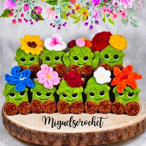 May include: A group of green crochet creatures with smiling faces, decorated with colourful flower accents. The creatures have brown, round bases and are arranged on a wooden slice. The text "Miguels crochet" is at the bottom.