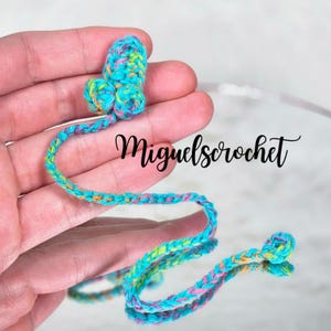 May include: A colourful crocheted bookmark with a flower-shaped top. The bookmark is made with pink, blue, green, and yellow yarn. The bookmark is held in a hand, and the text "miguelscrochet" is visible in the background.