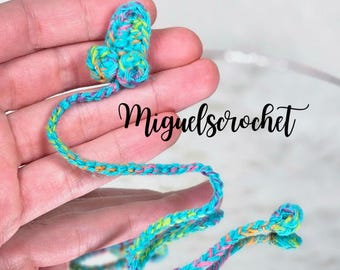 May include: A colorful crocheted bookmark with a flower-shaped top. The bookmark is made with pink, blue, green, and yellow yarn. The bookmark is held in a hand, and the text "miguelscrochet" is visible in the background.