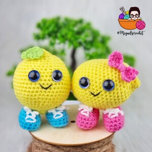 May include: Two crocheted lemon-shaped plushies with smiling faces, blue and pink shoes, and a pink bow on one. The plushies are sitting on a wooden surface.