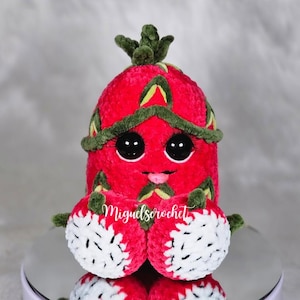 May include: A crocheted plush toy shaped like a strawberry with a green leafy top. The strawberry has large black eyes, a pink mouth, and white crocheted feet. The toy is sitting on a reflective surface.