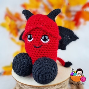 May include: A red crocheted devil plush toy with black horns, wings, and feet. The toy has a smiling face with white eyes and a black mouth.