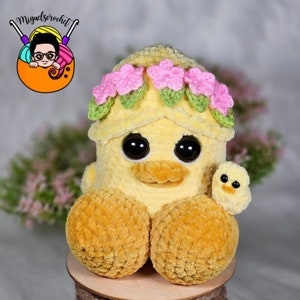 May include: A crocheted yellow chick wearing a pink flower crown and holding a smaller yellow chick. The chicks are sitting on a wooden surface.