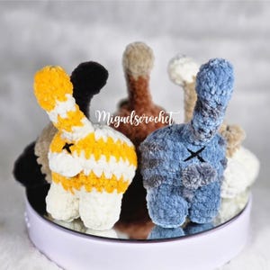 May include: A selection of handmade crochet bunny butt plushies in various colours, including yellow and white, blue, black, and brown. Each plush features a tail and a stitched "X" detail. The text "Miguels crochet" is visible.