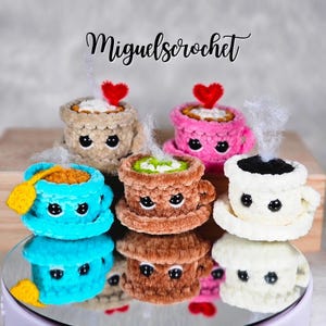 以下が含まれることがあります： ベージュ、ピンク、ブルー、ブラウン、ホワイトなど、様々な色の編みぐるみのコーヒーカップとティーカップのコレクション。各カップには笑顔の顔があり、上部から湯気が立ち上っています。一部のカップにはハートが乗っています。「Miguels crochet」の文字が見えます。