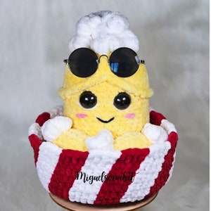 May include: A crocheted popcorn bucket with a smiling face, wearing sunglasses and a white crocheted hat. The bucket is filled with white crocheted popcorn and has red and white stripes.