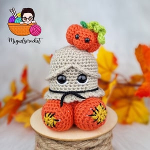May include: A crocheted pumpkin character with a small pumpkin on its head. The character has large black eyes and is wearing a beige hat with a black ribbon. The character is sitting on a wooden spool of twine.