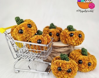 Op de afbeelding: Een miniatuur winkelwagentje gevuld met zes gehaakte oranje pompoenen met zwarte ogen. De pompoenen staan op een wit oppervlak met een bruin touw achter de kar. De tekst '@Migueltscrochet' is zichtbaar op de achtergrond.