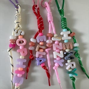 May include: Four personalised keychains featuring colourful paracord lanyards with plastic lobster clasps. Each keychain displays a name spelled out in block letter beads, including SOPHIE, ELLA, IZZY, and HENRY, interspersed with various round beads.
