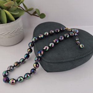 May include: A faceted bead necklace with iridescent colors of purple, green, and blue. The necklace is strung on a dark cord and has a gold-colored clasp. The necklace is displayed on a dark gray, heart-shaped jewelry box.