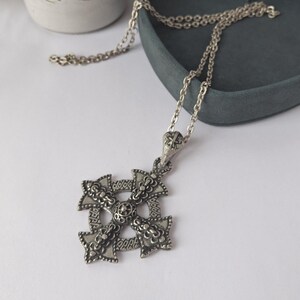 May include: Silver-toned pendant necklace featuring a detailed, ornate cross design. The cross has intricate patterns and a textured surface, suspended from a delicate chain. The necklace is displayed on a white surface with a dark gray tray in the background.