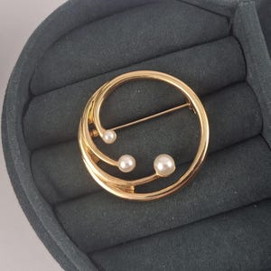 May include: A gold-coloured brooch with a circular design, featuring three faux pearls and a pin closure. The brooch has a modern, minimalist aesthetic, suitable for adding a touch of elegance to any outfit. The brooch is displayed on a dark grey velvet surface.