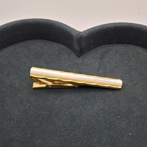 May include: A gold-toned metal tie bar featuring a central horizontal inlay of smooth white material. The tie bar has a textured, faceted edge design along the sides and a spring-loaded clip mechanism on the back for securing to a necktie.