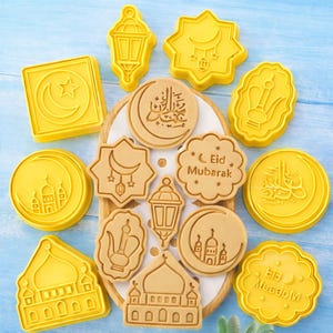 May include: Yellow and tan cookie cutters in various shapes, including a crescent moon and star, a mosque, and a lantern. Some cutters have Arabic script and the words "Eid Mubarak". The cutters are arranged on a wooden tray.