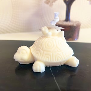 May include: A white ceramic turtle candle with a smaller turtle on its back. The candle is detailed with a textured shell and has a white wick.